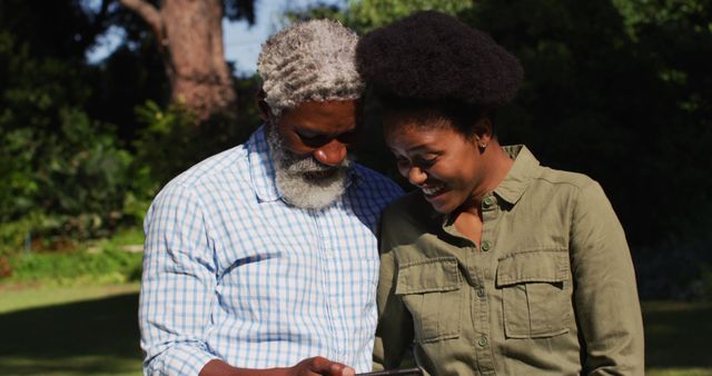 Mature couple happily sharing a moment while looking at a tablet in a lush, green garden. Afro hair woman and man with grey beard seem connected and engaged, ideal for use in advertisements or articles about elder tech adoption, enjoying retirement, or family tech trends.