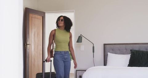 African american woman entering modern hotel bedroom pulling suitcase and carrying bag