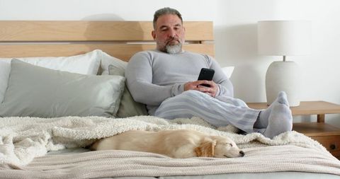 Mature man relaxing in bed holding smartphone with sleeping puppy, cozy neutral bedroom