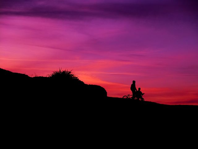 Image depicts two people on bikes silhouetted against a vibrant purple and pink sunset sky. Perfect for themes of adventure, outdoor activities, family bonding, or promoting cycling events.