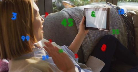 Chinese woman waving and using tablet on couch at home with colorful numeric overlay