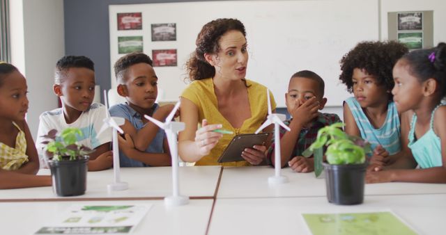 A teacher is teaching a diverse group of young children about renewable energy sources in a classroom. The children are attentively listening while seated around a table that has wind turbine models and plants. This image can be used for educational content, school websites, STEM program promotions, sustainability education materials, and eco-friendly initiatives targeting young learners.