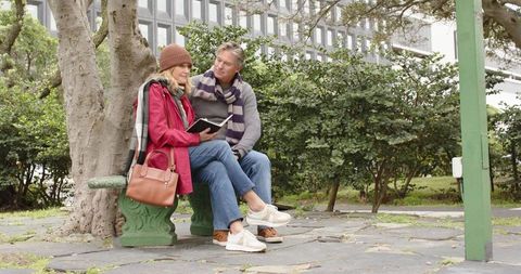 Middle-aged couple sitting on green bench in city park reading notebook and smiling
