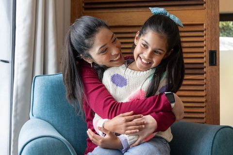 Mother hugging daughter on teal armchair showing smartwatch, cozy home family bonding moment
