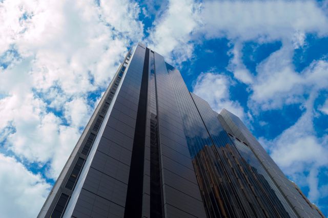 Photo of a towering modern skyscraper with a glass facade reflecting a blue sky and white clouds. This image is ideal for projects related to architecture, urban development, corporate environments, and financial or commercial themes. It invokes a sense of modernity, progress, and professionalism, making it suitable for websites, presentations, advertisements, and promotional materials for businesses or real estate developments.