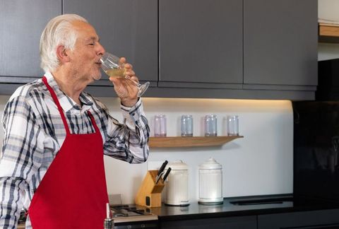 Senior man sipping white wine in modern kitchen wearing red apron and plaid shirt