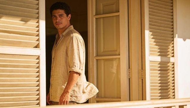 Man standing on sunlit veranda wearing linen shirt gazing through open louvered shutters