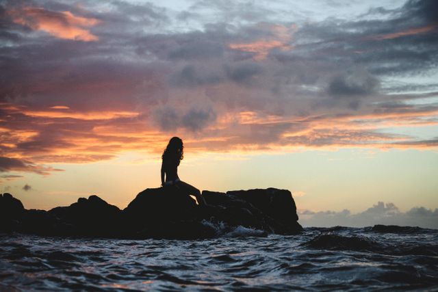 This scene captures the serene beauty of a sunset over the ocean. A woman sits peacefully on rocks, silhouetted against the vibrant colors of the sky. Ideal for use in travel promotions, wellness and meditation themes, or any nature-related advertisement. The dramatic clouds and the gentle waves add to the tranquility and majestic feel of the moment.