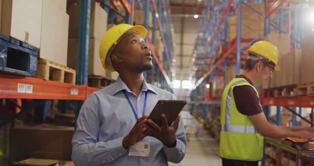 African american male worker with helmet using tablet in warehouse. global business, shipping and delivery.