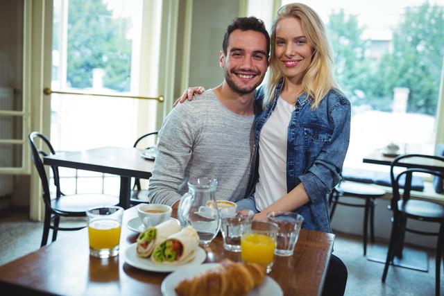 Young couple sitting together in a cozy café, smiling and enjoying breakfast. The table is set with orange juice, coffee, water, croissants, and wraps. Ideal for use in lifestyle blogs, relationship articles, café promotions, and social media content highlighting happy moments and dining experiences.