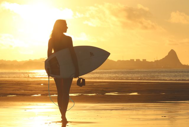 Young woman carrying surfboard along beach during sunset, creating striking silhouette against golden sky. Perfect for themes of active lifestyle, summer vacations, beach activities, surfing culture, and oceanic leisure. Great for travel websites, sport promotions, advertisements for beachwear.
