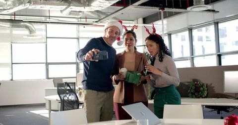 Multicultural office team posing for festive selfie while exchanging gifts and wearing holiday hats
