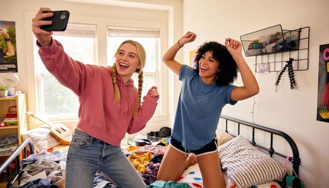 Friends taking selfie and dancing on unmade bed in bright casual bedroom morning