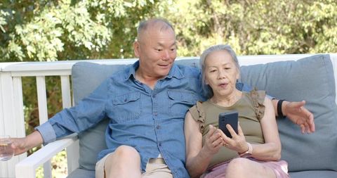 Senior asian couple relaxing on patio sofa using smartphone and sharing drink