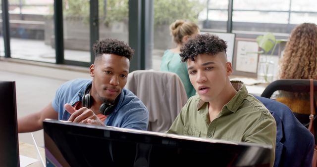 Two young men are collaborating at a computer in a modern office. They appear engaged and are discussing something on the screen. The background features large windows, a spacious and light-filled environment, and other colleagues working. Great for illustrating teamwork, modern work environments, collaborative efforts, and diverse professional settings. Suitable for use in articles or promotions about modern workplaces, team collaboration, tech industry, and professional development.