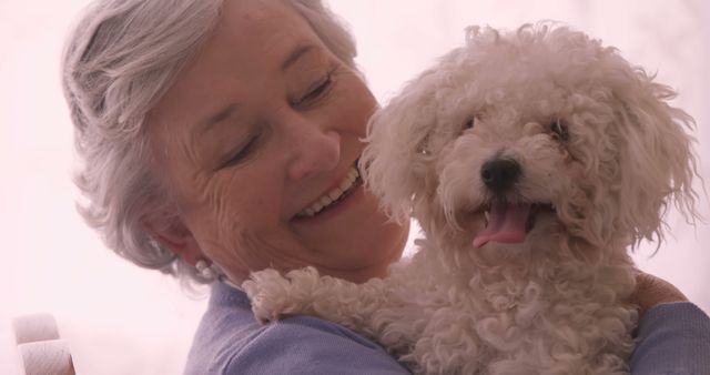 A senior Caucasian woman is affectionately holding a small white dog, both appearing joyful and content. Their bond exemplifies the companionship and happiness pets can bring to the lives of elderly individuals.
