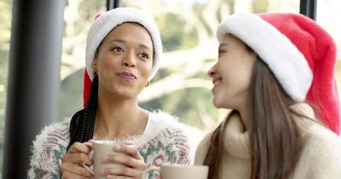 Friends enjoying cozy holiday coffee wearing santa hats by large window
