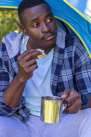 African american camper eating sandwich and holding metal mug at tent entrance