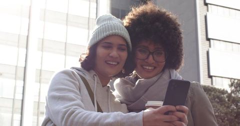 Friends sharing coffee while checking smartphone on sunlit urban street