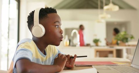 African american boy wearing headphones studying online at home desk with laptop and pencil