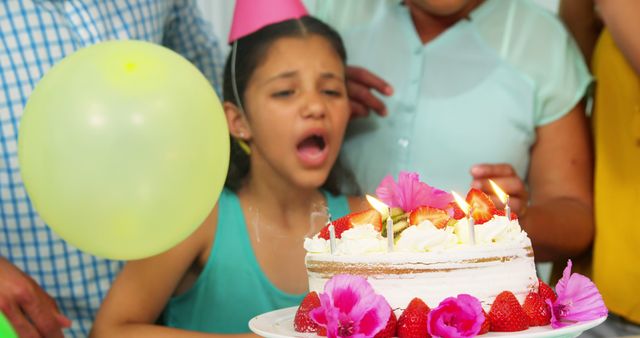 Young girl blowing out candles on her birthday cake, surrounded by family and friends. Ideal for use in articles or advertisements about children's parties, birthday celebrations, family gatherings, and special occasions. Can be used to promote birthday supplies, party planning services, or feature ideas for hosting kids' birthday parties.
