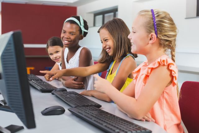 Group of diverse schoolgirls enjoying time on computers in a classroom. They are smiling and pointing at the screen, indicating a fun and interactive learning environment. This image can be used for educational content, school promotions, technology in education, and children's activities.
