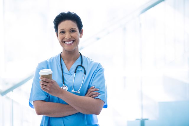 This image shows a smiling female nurse holding a disposable coffee cup in a hospital corridor. She is wearing blue scrubs and a stethoscope around her neck, indicating she is a healthcare professional. This image can be used for healthcare-related content, promoting nursing careers, hospital advertisements, or articles about healthcare workers' daily lives.