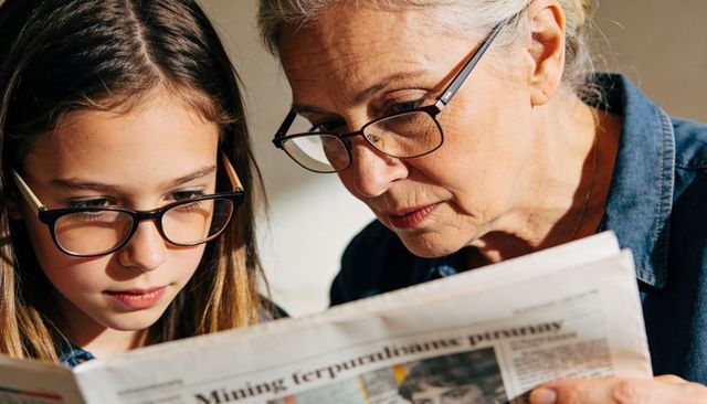 Senior woman teaching granddaughter to read newspaper at home, closeup eyewear moment