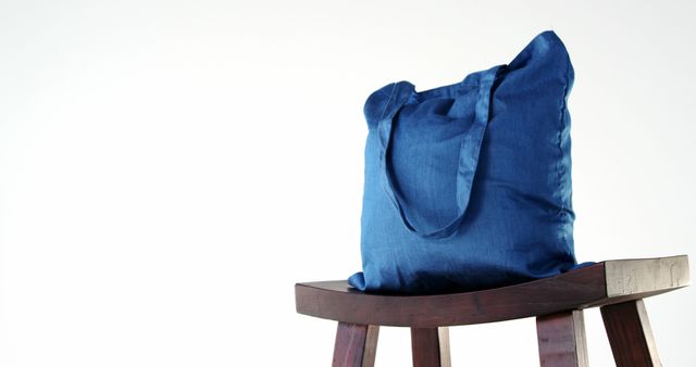 A blue tote bag rests on a wooden stool against a white background, with copy space. Its simplicity and the contrast of colors make it ideal for showcasing the design of everyday accessories.