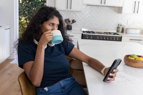 African american woman relaxing at kitchen island drinking coffee and checking smartphone