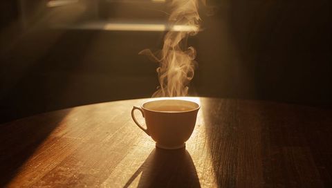 Steaming ceramic cup on sunlit wooden table casting long shadow, cozy morning mood