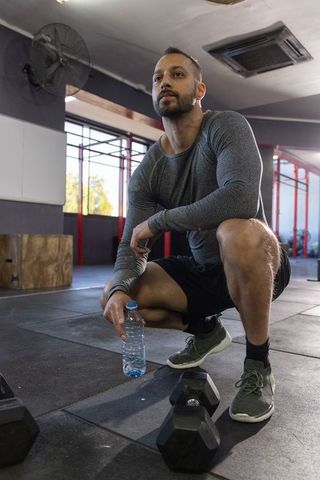 Mid-adult indian man crouching in gym holding water bottle by dumbbells