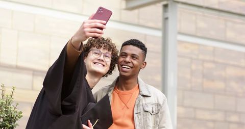 Young male friends taking graduation selfie on campus, smiling celebrating achievement
