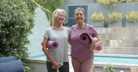 Mature women carrying yoga mats and water bottles on sunny backyard pool patio
