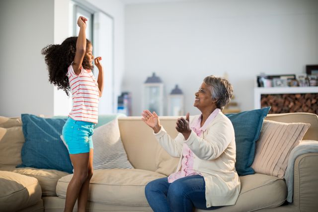 Grandmother applauding her granddaughter while she is dancing in the living room. This image captures a joyful family moment, highlighting the bond between generations. Perfect for use in family-oriented advertisements, articles on family relationships, or promotions for home and lifestyle products.