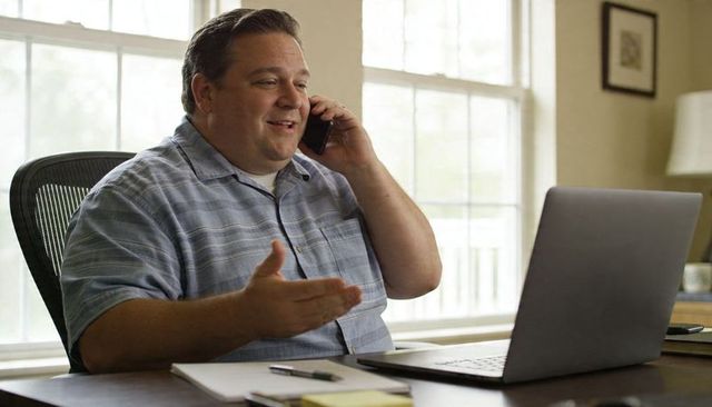 Smiling man talking on phone and gesturing during remote work at sunlit home office with laptop