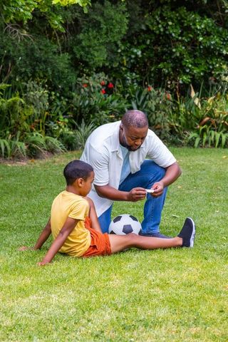 African american father tending son's leg after soccer injury in backyard garden