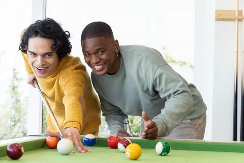Multicultural friends playing pool in sunlit game room laughing during casual leisure