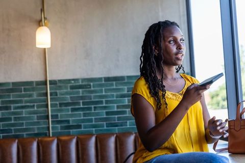 African woman looking out window holding smartphone in sunlit cafe wearing mustard blouse