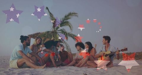 Friends sitting on beach at dusk listening to man playing guitar holding string lights