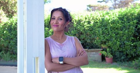 Confident indian woman leaning on white porch post wearing smartwatch in sunlit garden