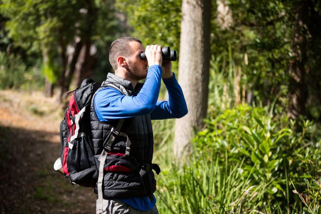 Male hiker with a backpack using binoculars to observe nature in a lush forest. Ideal for content related to outdoor activities, adventure travel, wildlife observation, and nature exploration.