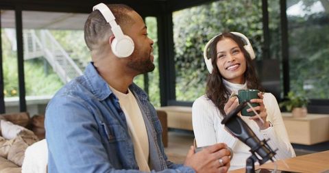Diverse podcast cohosts recording with white headphones and tabletop mic in modern studio