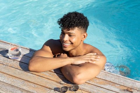 Smiling man relaxing at pool edge leaning on wooden deck with sunglasses and citrus drink