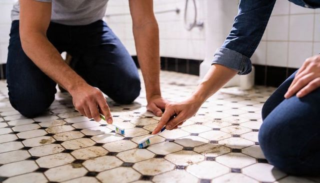 Couple scrubbing grimy hex tile grout with toothbrushes kneeling in bathroom