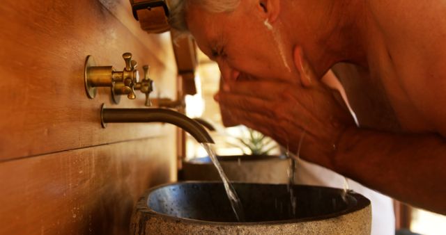 A senior Caucasian man is washing his face at a sink with an antique brass faucet, with copy space. Capturing a moment of personal hygiene, the image reflects a routine aspect of daily life.