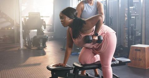 African american woman performing dumbbell row with female trainer guiding form in gym