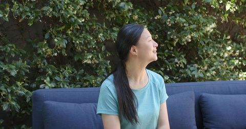 Asian woman relaxing on navy sofa looking upward at sunlit garden vines and foliage