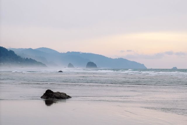 Featuring a serene coastal morning with misty mountains lining the horizon and gentle waves lapping at the shore, this image evokes tranquility and calmness. Ideal for use in travel blogs, nature articles, and calming backgrounds, it depicts the natural beauty of the sea at dawn.