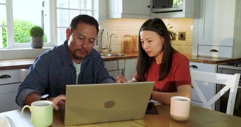 Asian couple working together on laptop in bright cozy kitchen, home office collaboration